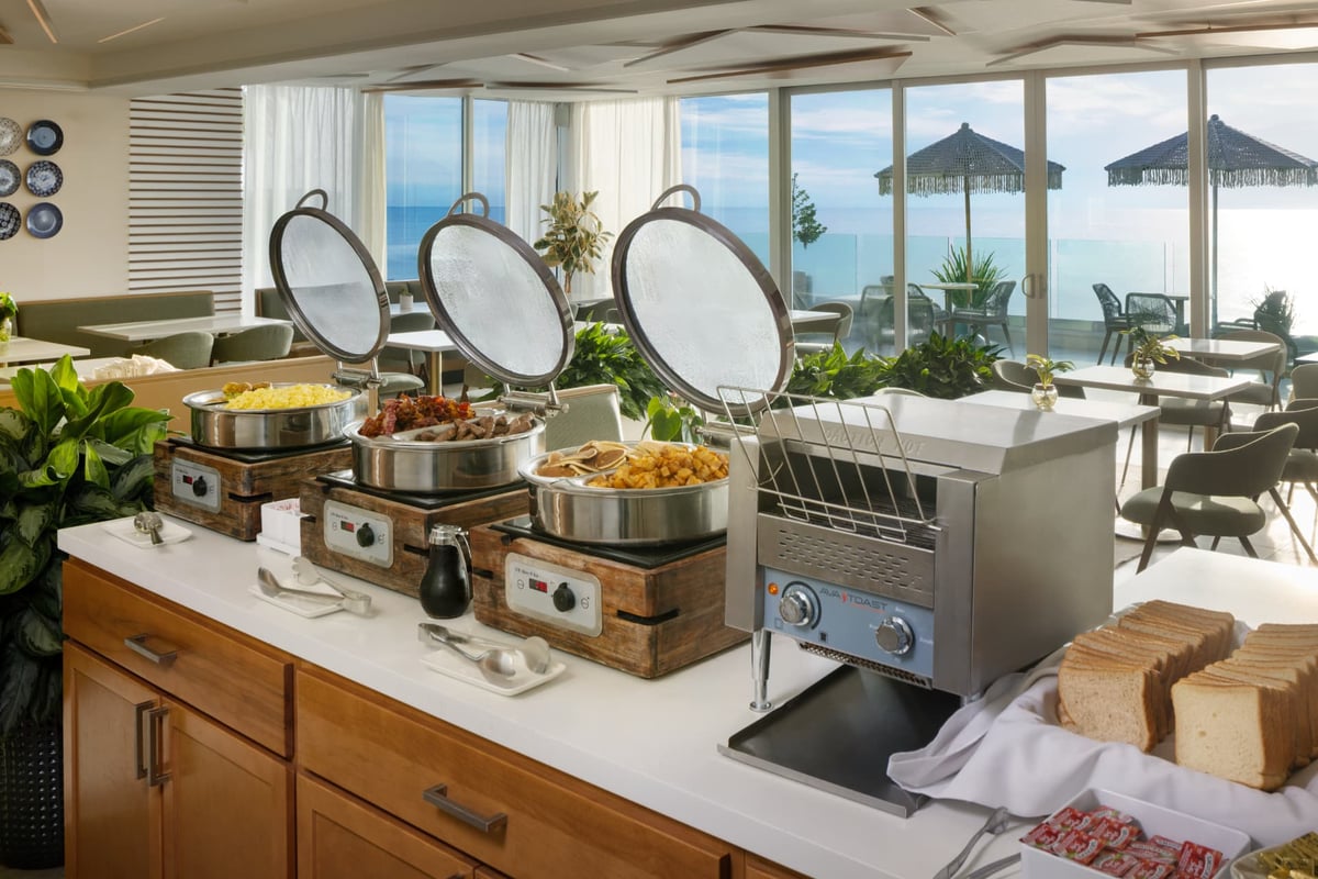 The breakfast service at MB Hotel — chafing dishes of eggs, bacon and potatoes along a white counter beside the toaster and bread station, the ocean and fringed umbrellas through the windows behind