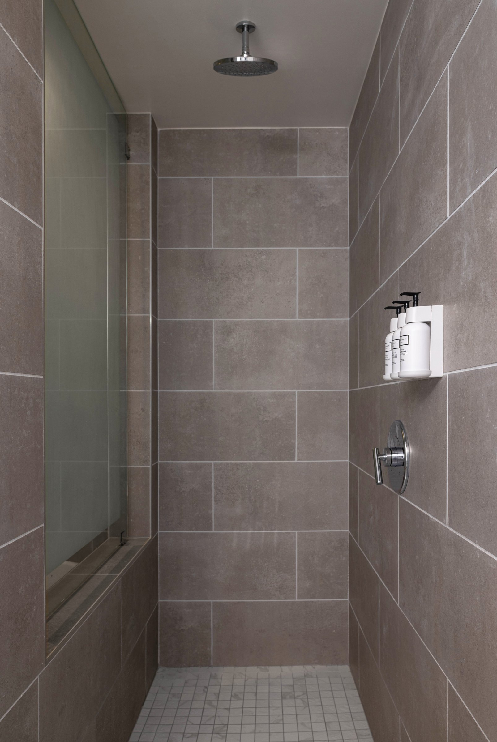 NK2 King with Jacuzzi — walk-in shower lined in warm grey tile with a ceiling-mounted rain head and wall-mounted amenity dispensers
