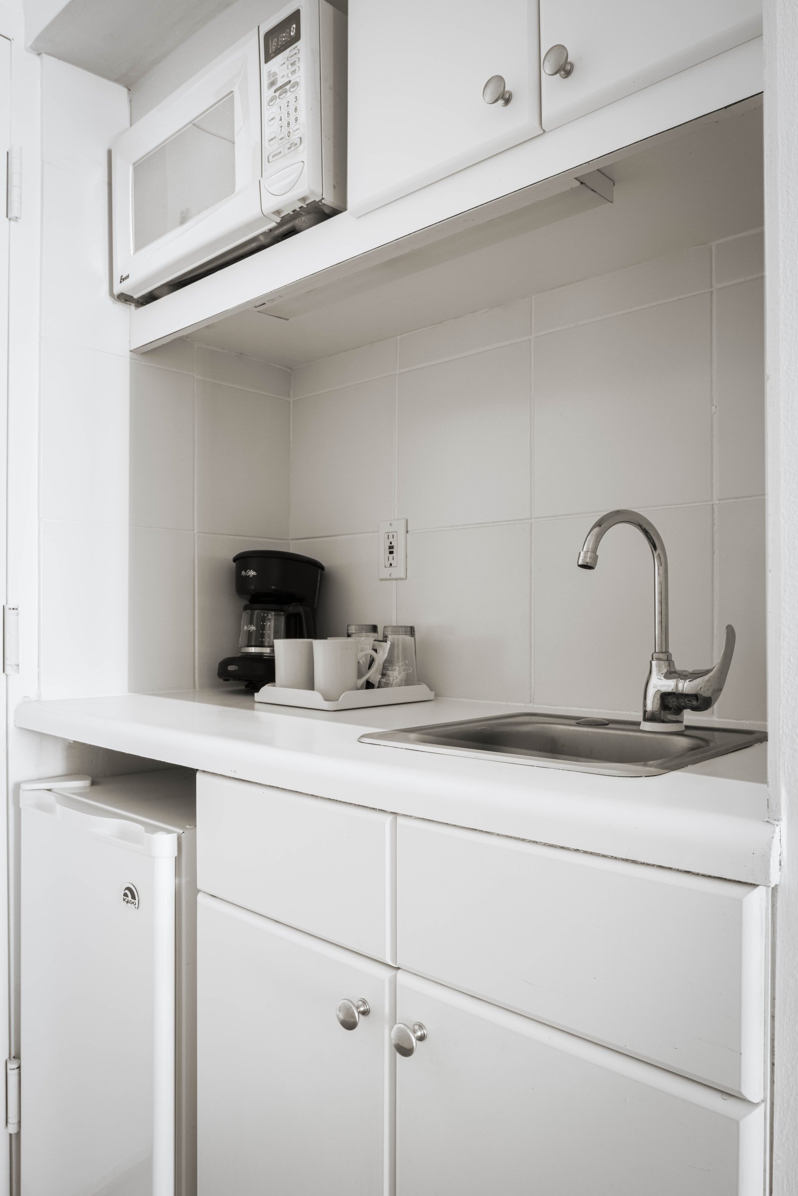 NK4 kitchenette — white cabinetry with an over-counter microwave, stainless sink, coffee maker on a tray and a small under-counter refrigerator