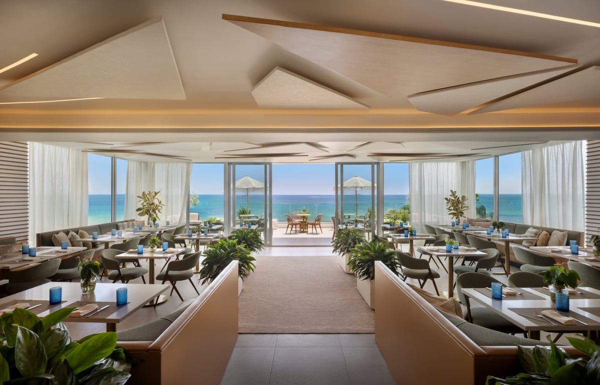 Kalypso dining room at midday, a full sweep of laid tables and planters beneath the sculpted ceiling, ocean on the horizon
