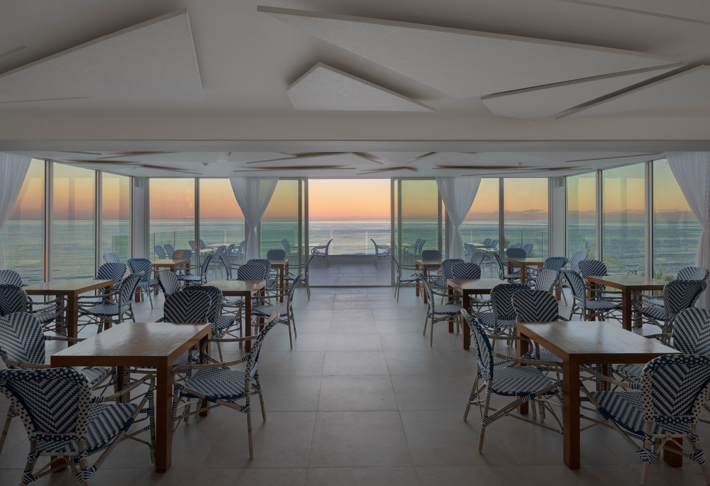 MB Hotel oceanfront event room at sunset — rows of blue-and-white bistro chairs facing floor-to-ceiling windows over the Atlantic
