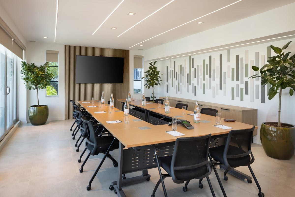 MB Hotel's larger boardroom, a long oak table under recessed light lines with a wall-mounted screen on a wood-slat wall and a felt art panel running the length of the room