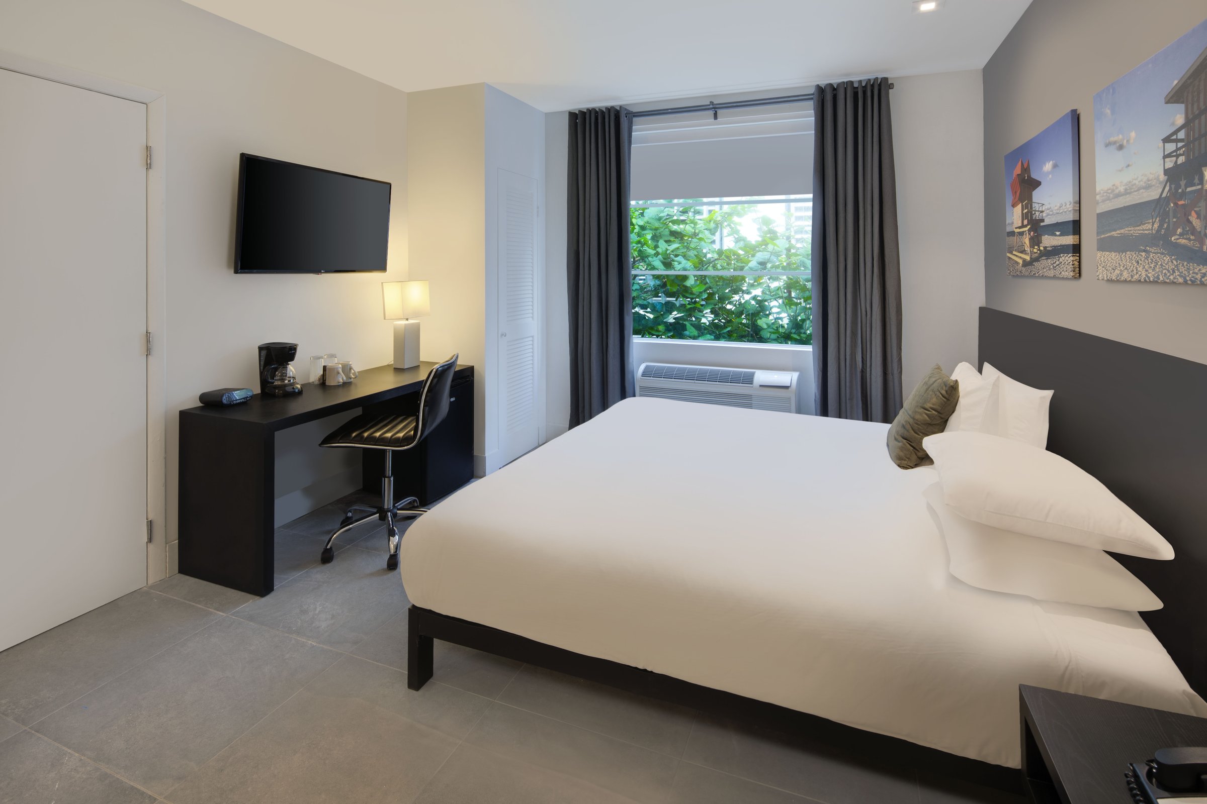 NK1 Standard King — king bed framed by a window of green sea-grape foliage, desk and mini fridge beside the entry door