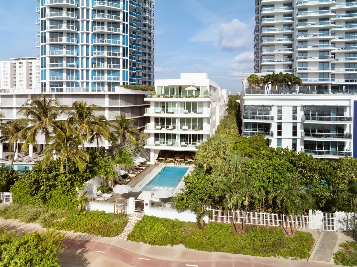 MB Hotel seen from above the beach — the pool deck and white balconied building set back from the Collins Avenue boardwalk, palms and sea grape between the two
