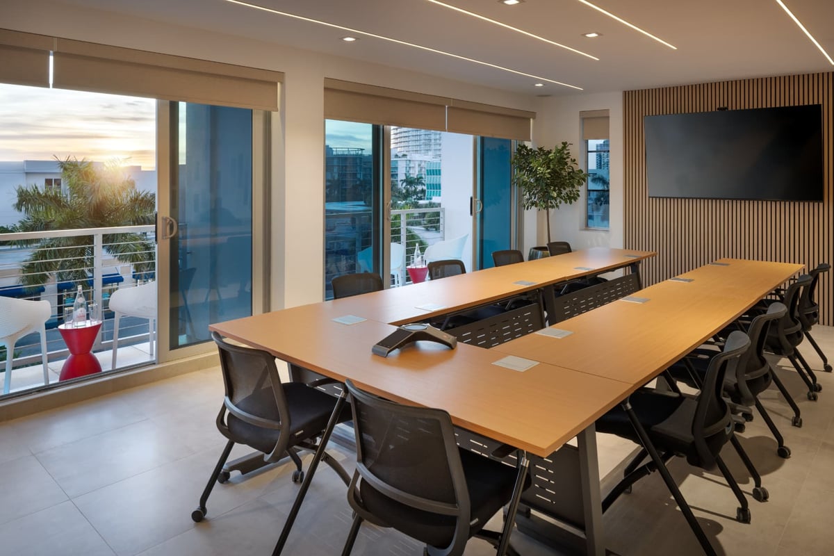 MB Hotel conference room, a long wood table with mesh-back chairs beside balcony doors opening to the ocean and the mid-Beach skyline