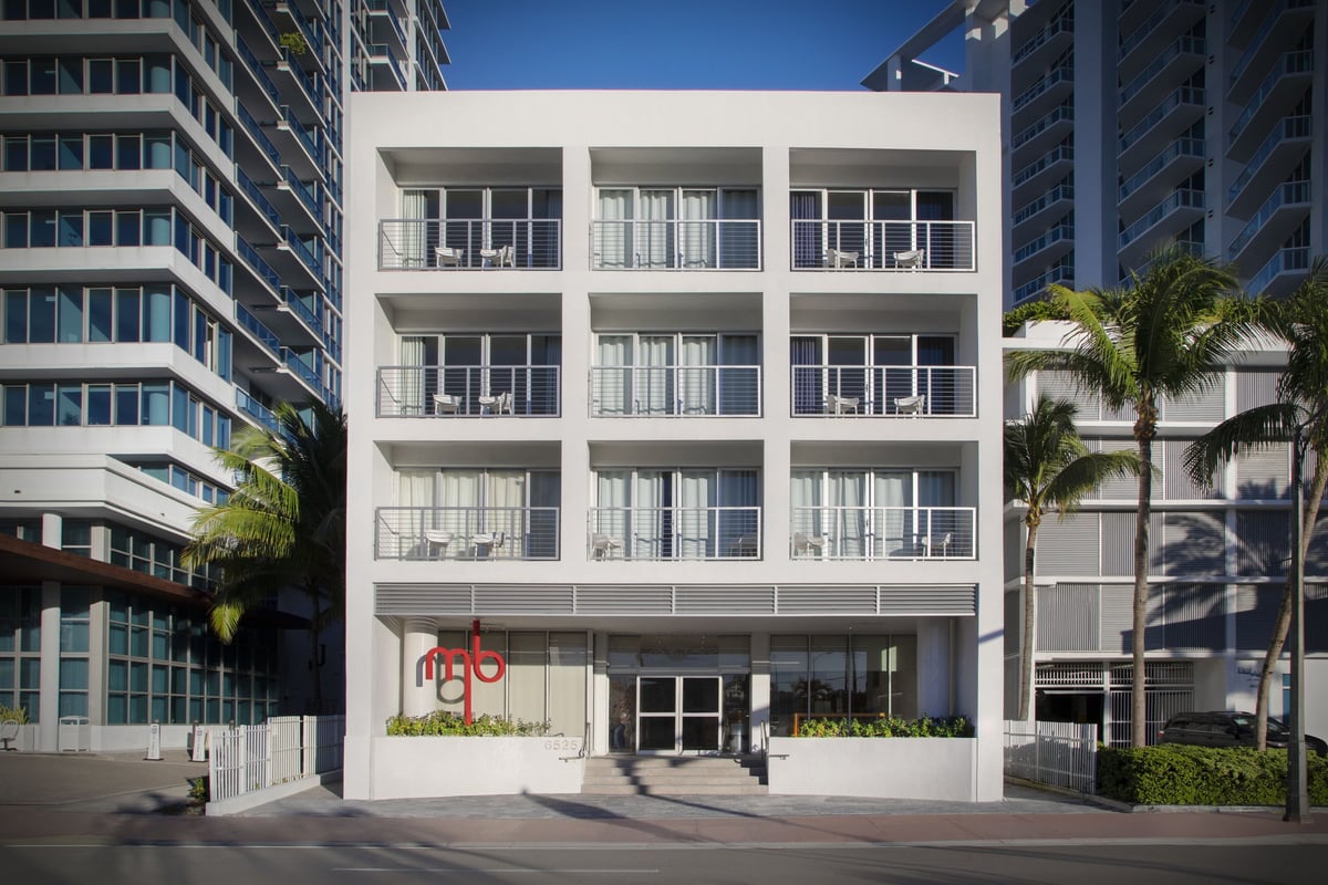 MB Hotel front facade by day — white balconied building on Collins Avenue with the red property mark above the entrance