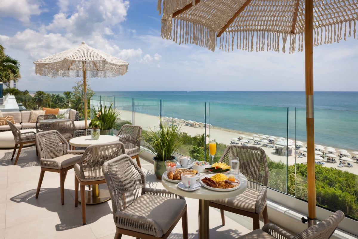 Breakfast on the MB Hotel rooftop terrace — pastries, fruit, cappuccino and a mimosa on a bistro table under a fringed macramé umbrella, the beach loungers and turquoise Atlantic below