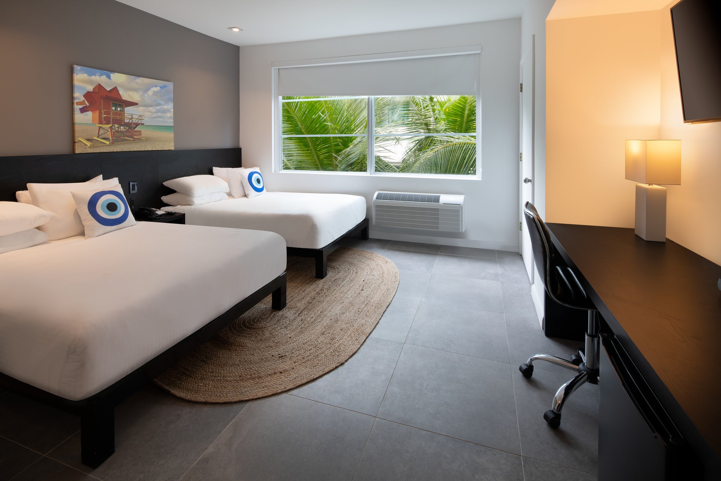 NDD1 Standard Double Full — the two beds seen from the desk wall, a round jute rug between them and palm fronds filling the window
