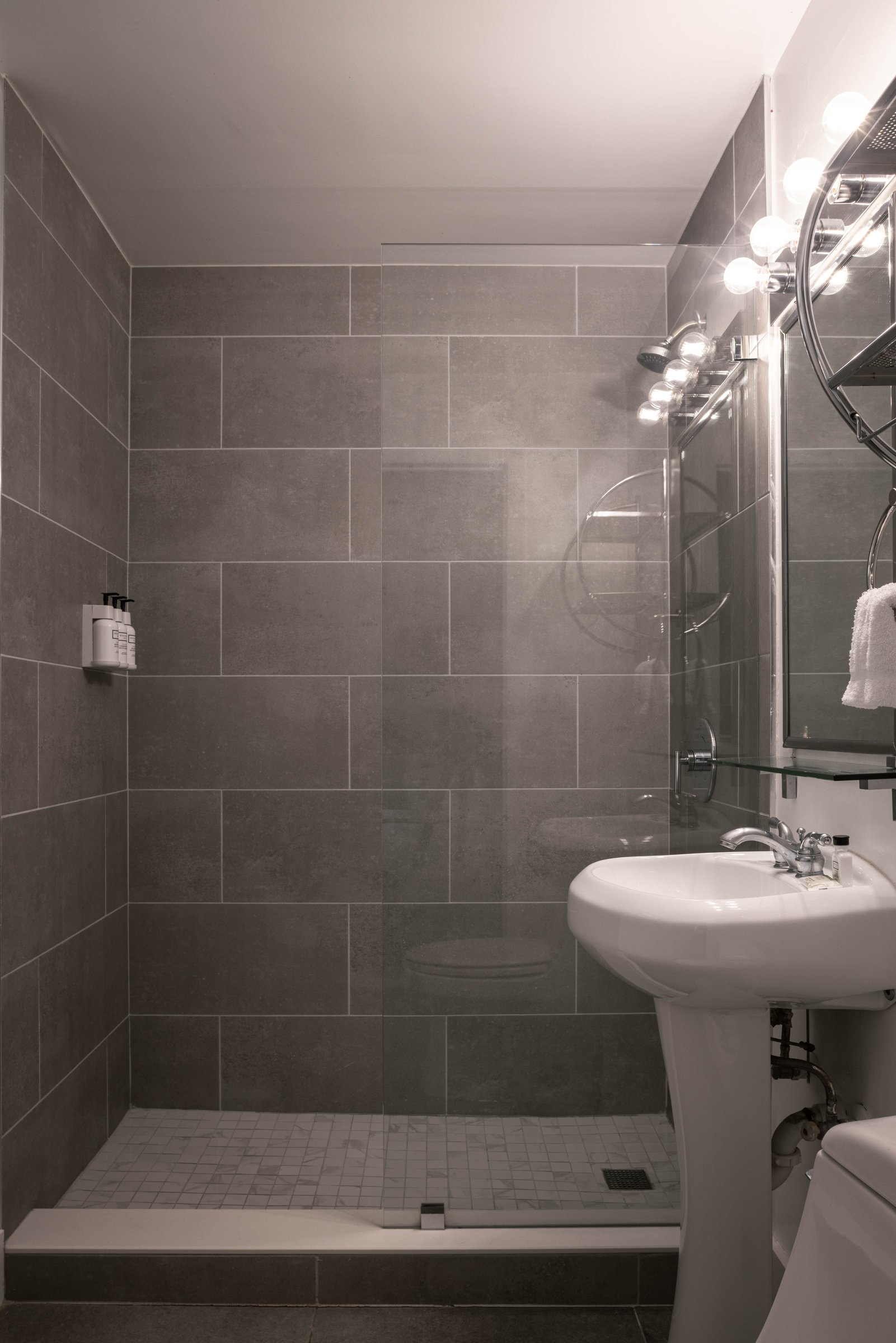 NK1 Standard King bathroom — grey stone-tiled walk-in shower behind a glass screen with a white pedestal basin and bulb-lit round mirror