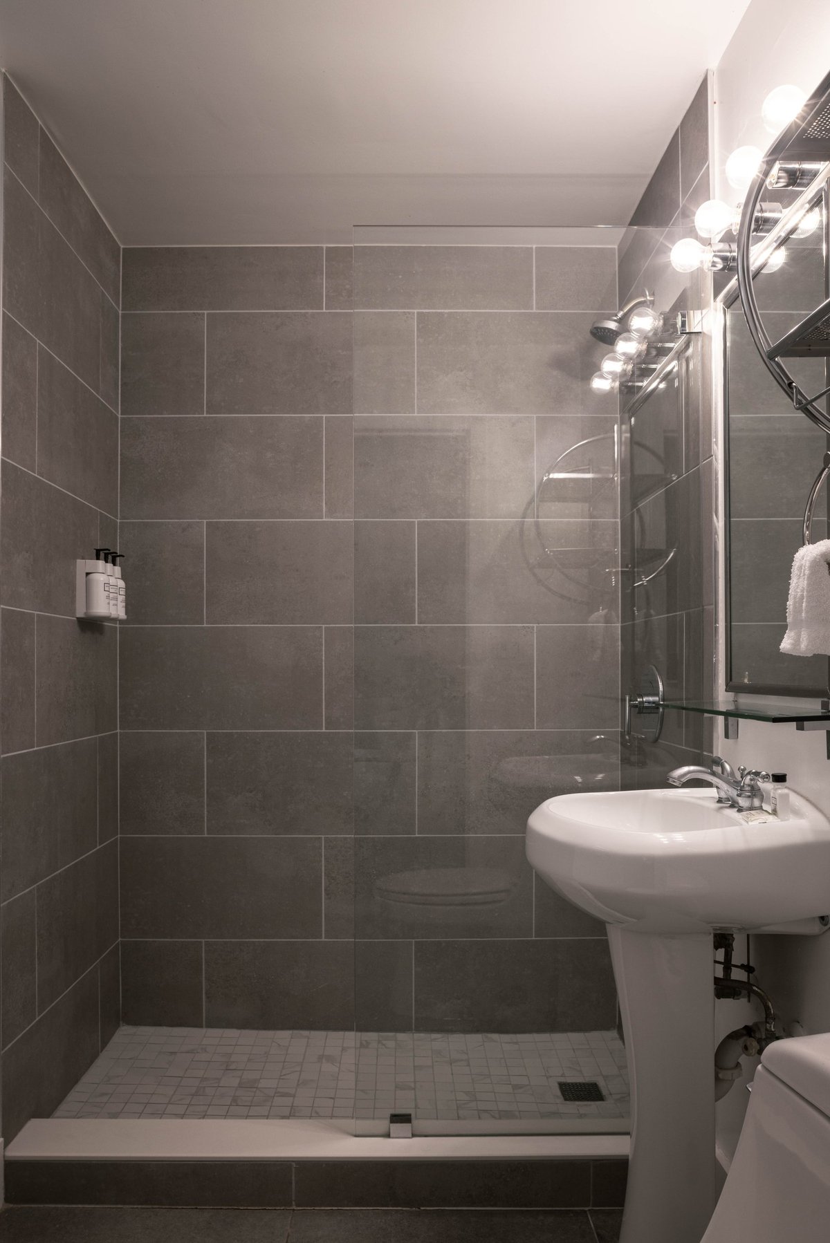 NK1 Standard King bathroom — grey stone-tiled walk-in shower behind a glass screen with a white pedestal basin and bulb-lit round mirror