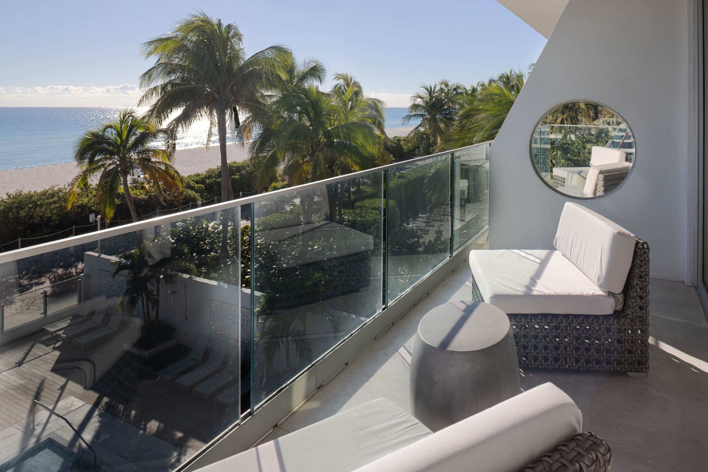 NK5 oceanfront balcony — glass balustrade above coconut palms with the beach and sunlit ocean beyond, cushioned wicker loungers and a concrete drum table on the terrace