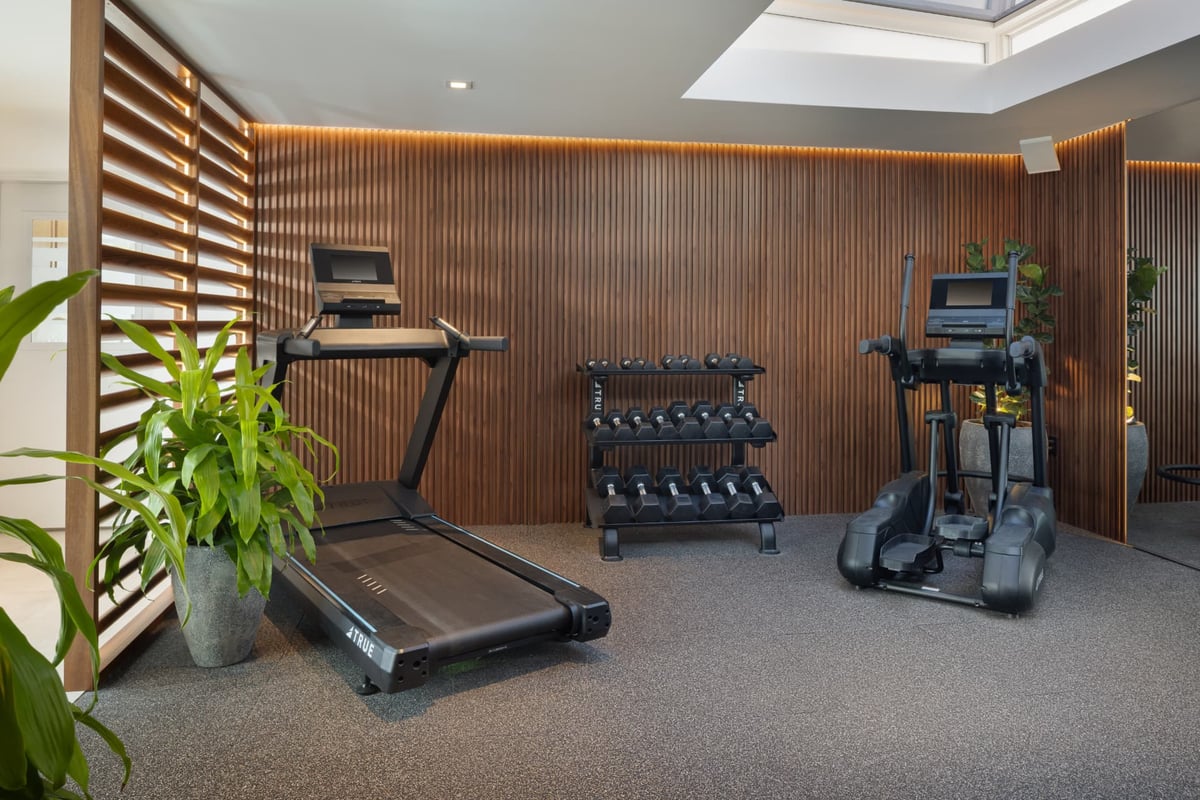 The MB Hotel fitness room — TRUE treadmill and elliptical, dumbbell rack and backlit walnut slat walls under a skylight