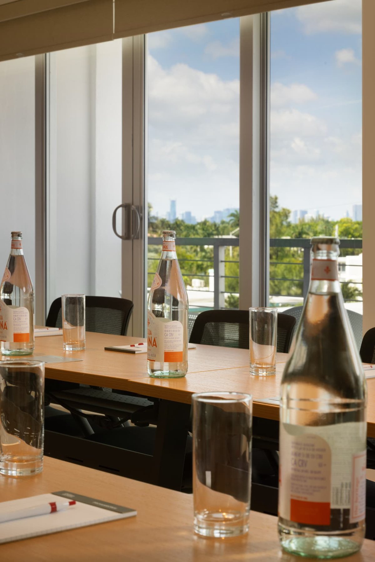 A meeting table dressed for a working lunch at MB Hotel — sparkling water, glassware and notepads along the oak top, the palms and downtown skyline through the balcony glass