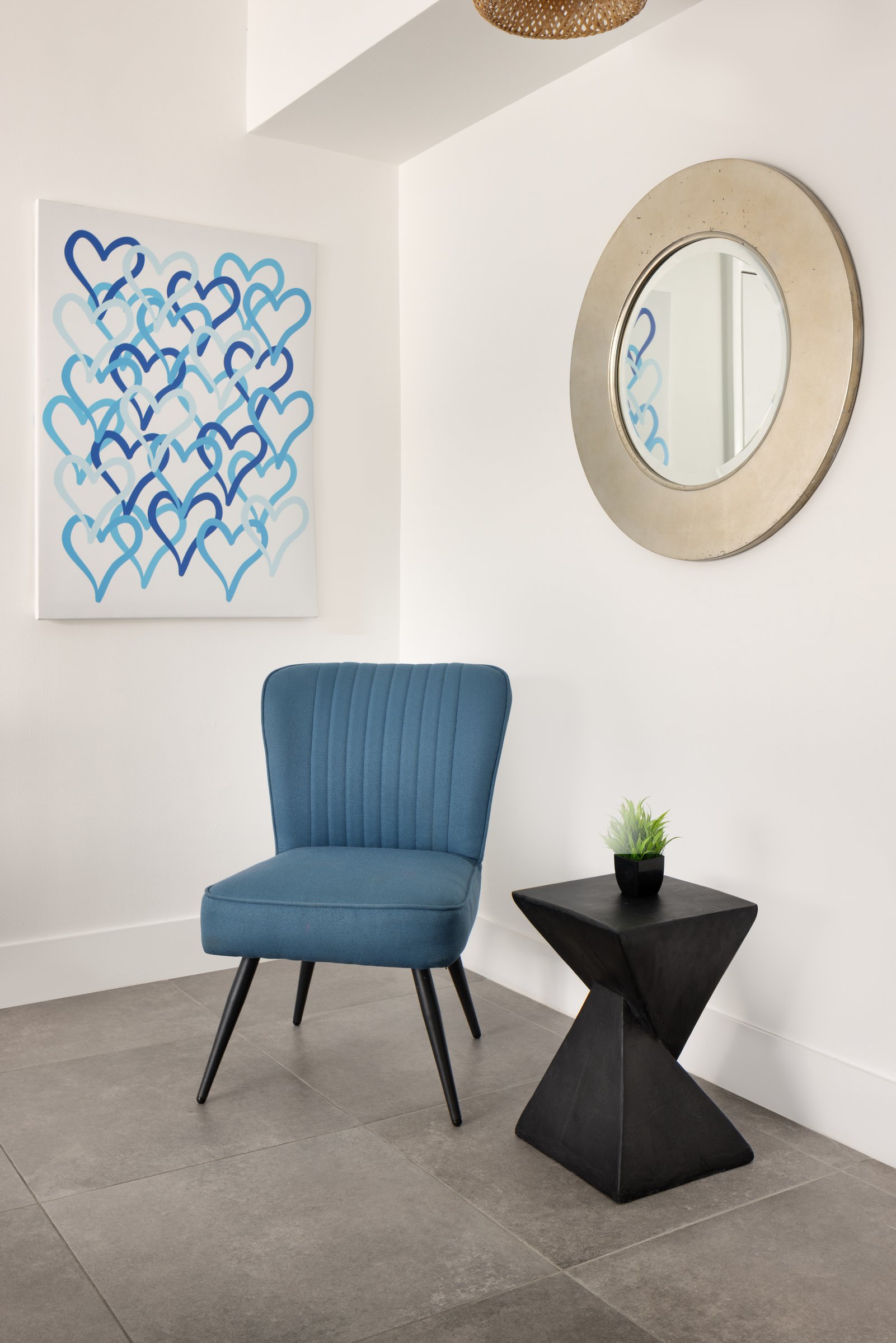 PNK1 entry corner — blue channel-backed chair beneath a canvas of overlapping blue hearts, with a round silver-framed mirror and a faceted black side table