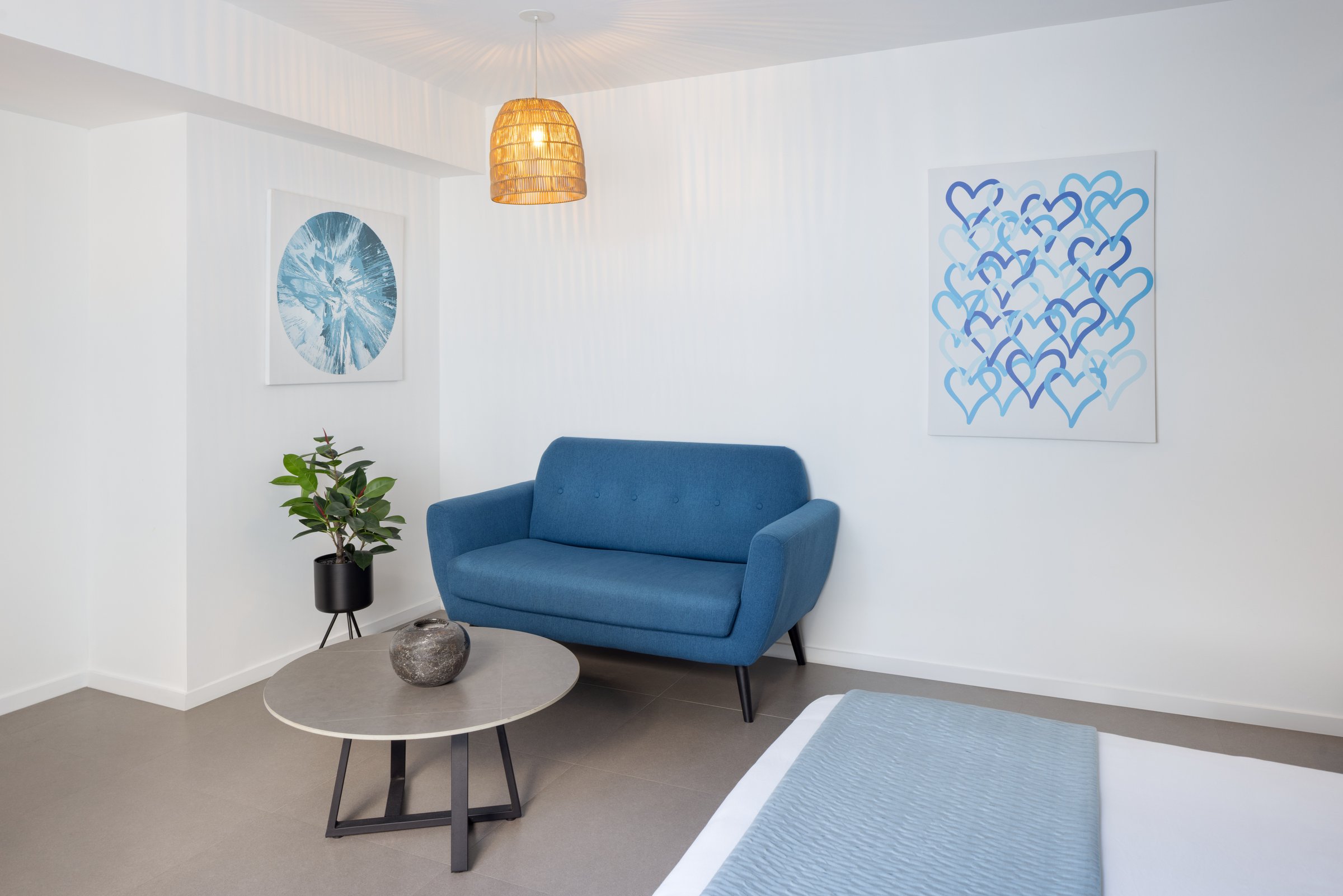 RM1 sitting area — blue two-seat sofa between two canvases, a potted plant and a round stone-topped coffee table beside the foot of the bed