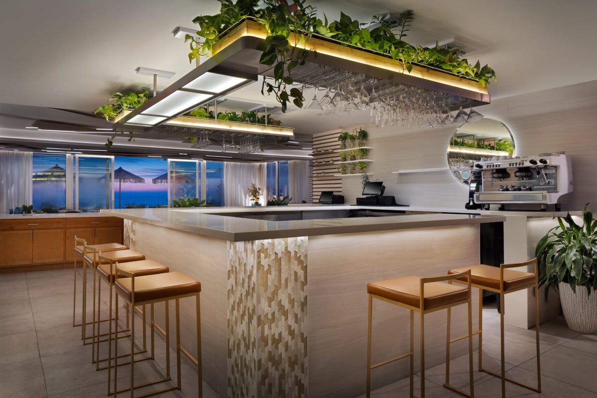 Kalypso's oceanfront bar at blue hour, glassware suspended beneath a planted light canopy with tan leather-and-brass stools along the counter and the beach umbrellas glowing outside