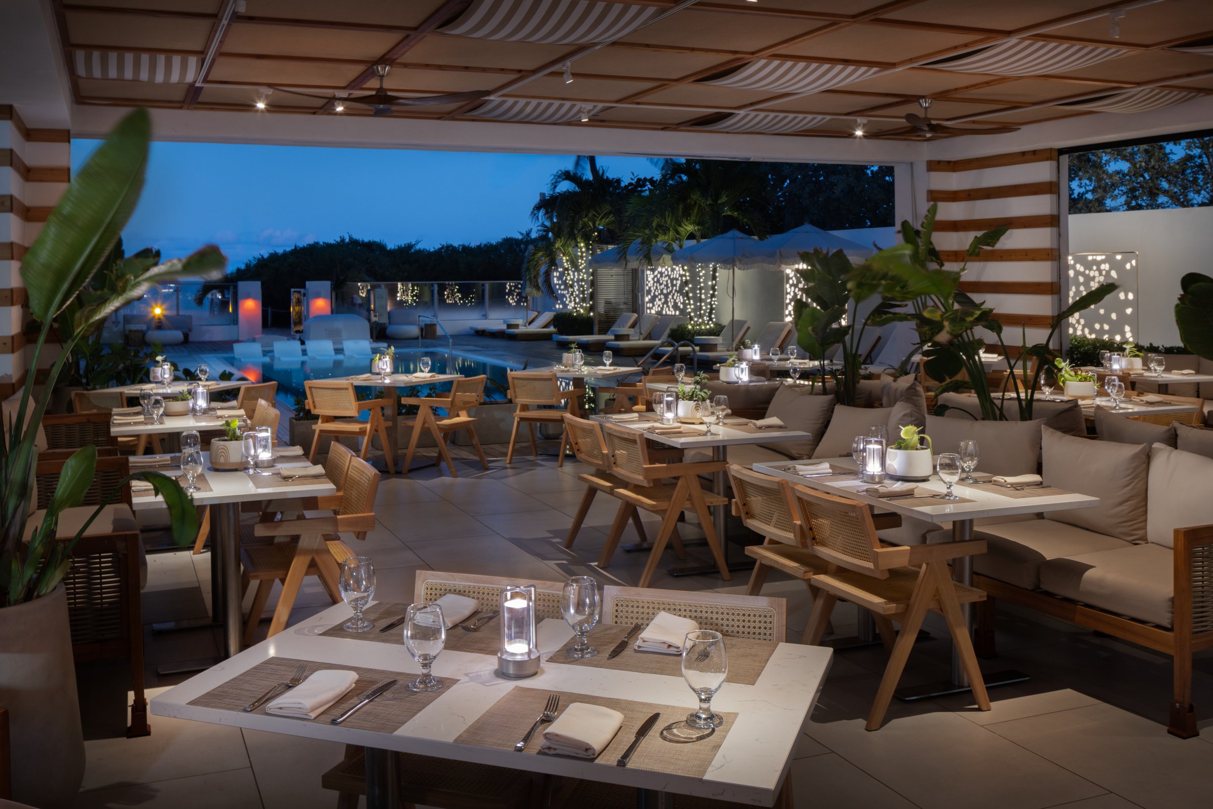 Kalypso terrace tables laid with linen and lanterns, the lit pool and cabanas in the background