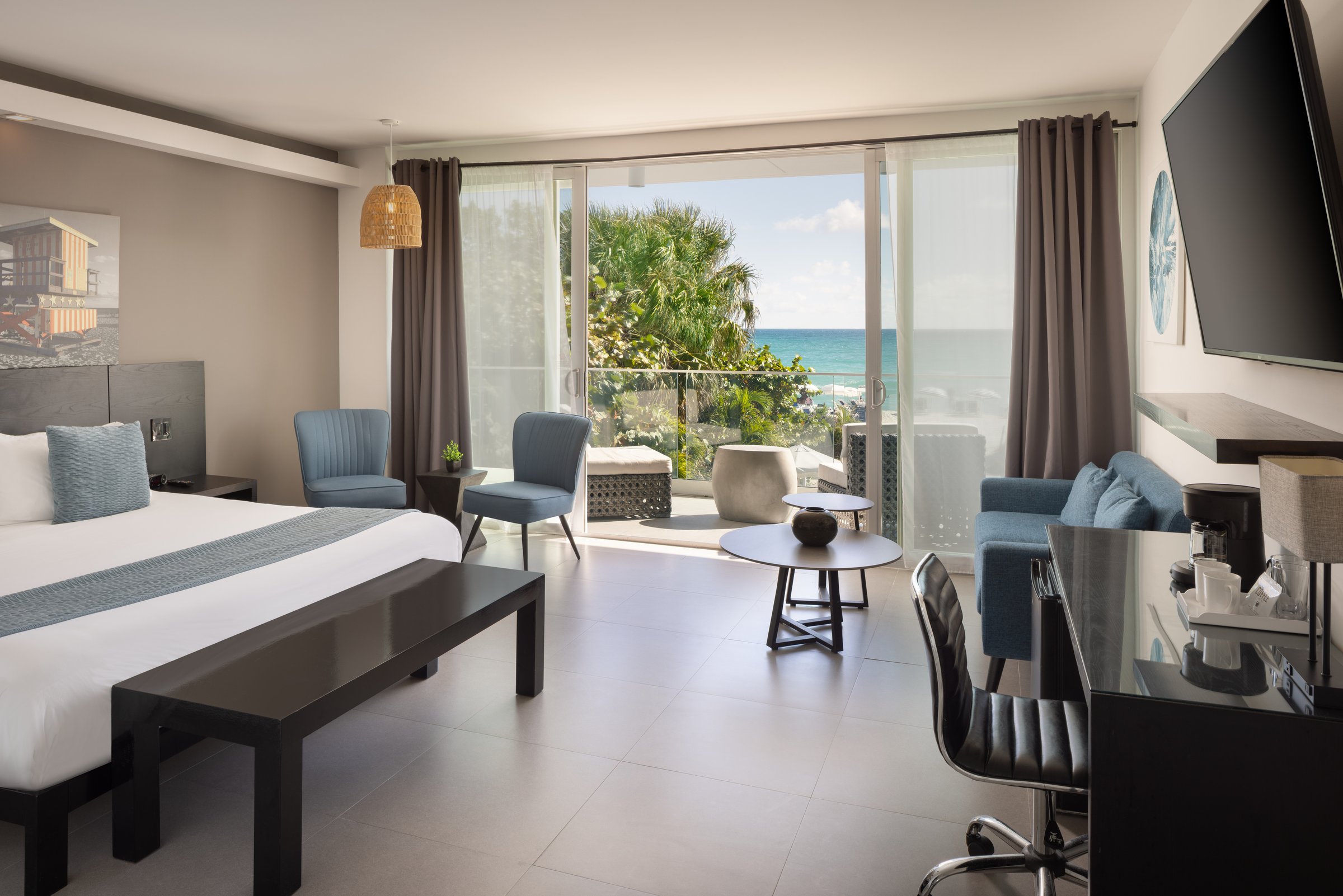 NK5 Deluxe Oceanfront King interior — king bed with a black bench at its foot, two blue chairs and a round table by the open balcony doors with the ocean visible past the palms