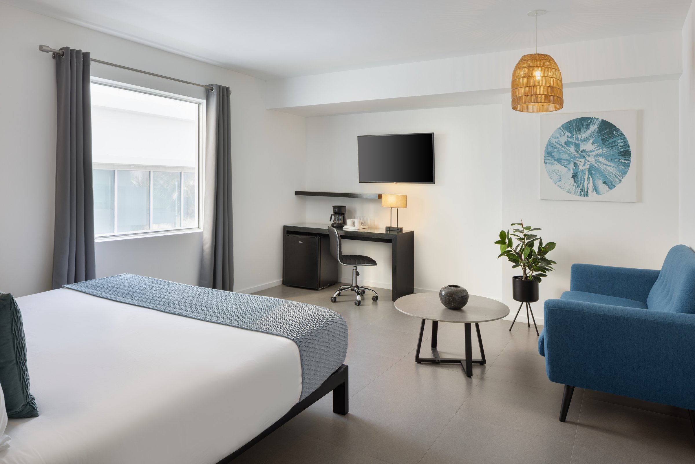 RM1 Deluxe King, Partial Ocean View — king bed with a pale blue runner, desk and television opposite, a blue sofa and round coffee table under a rattan pendant