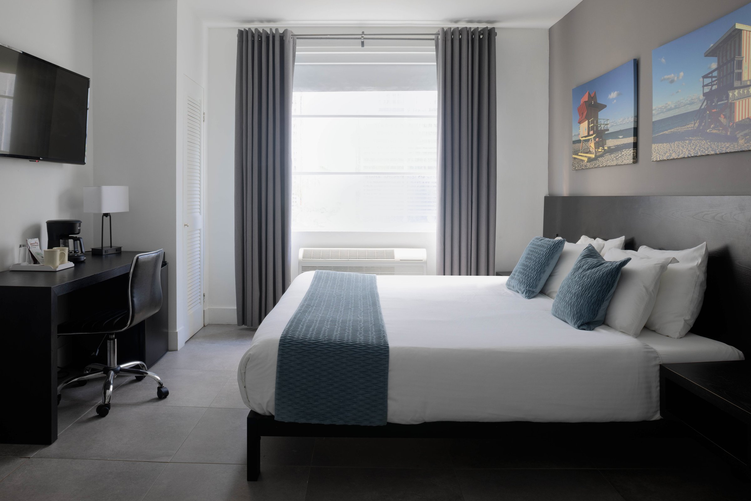 PNK1 Standard King, Accessible — king bed with a slate runner and blue cushions against a dark headboard, tall curtained window centre-room and a desk with coffee service opposite