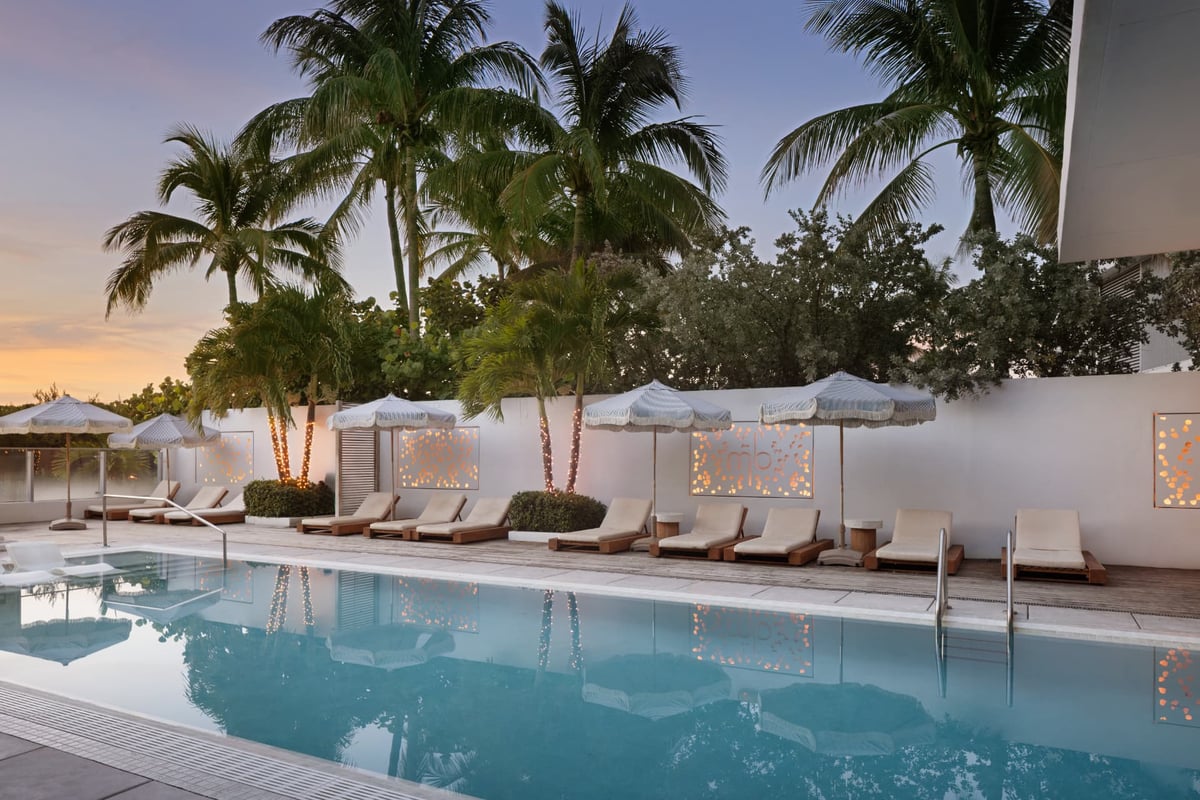 MB Hotel's pool deck at dusk, loungers arranged under palms with the illuminated mb monogram behind
