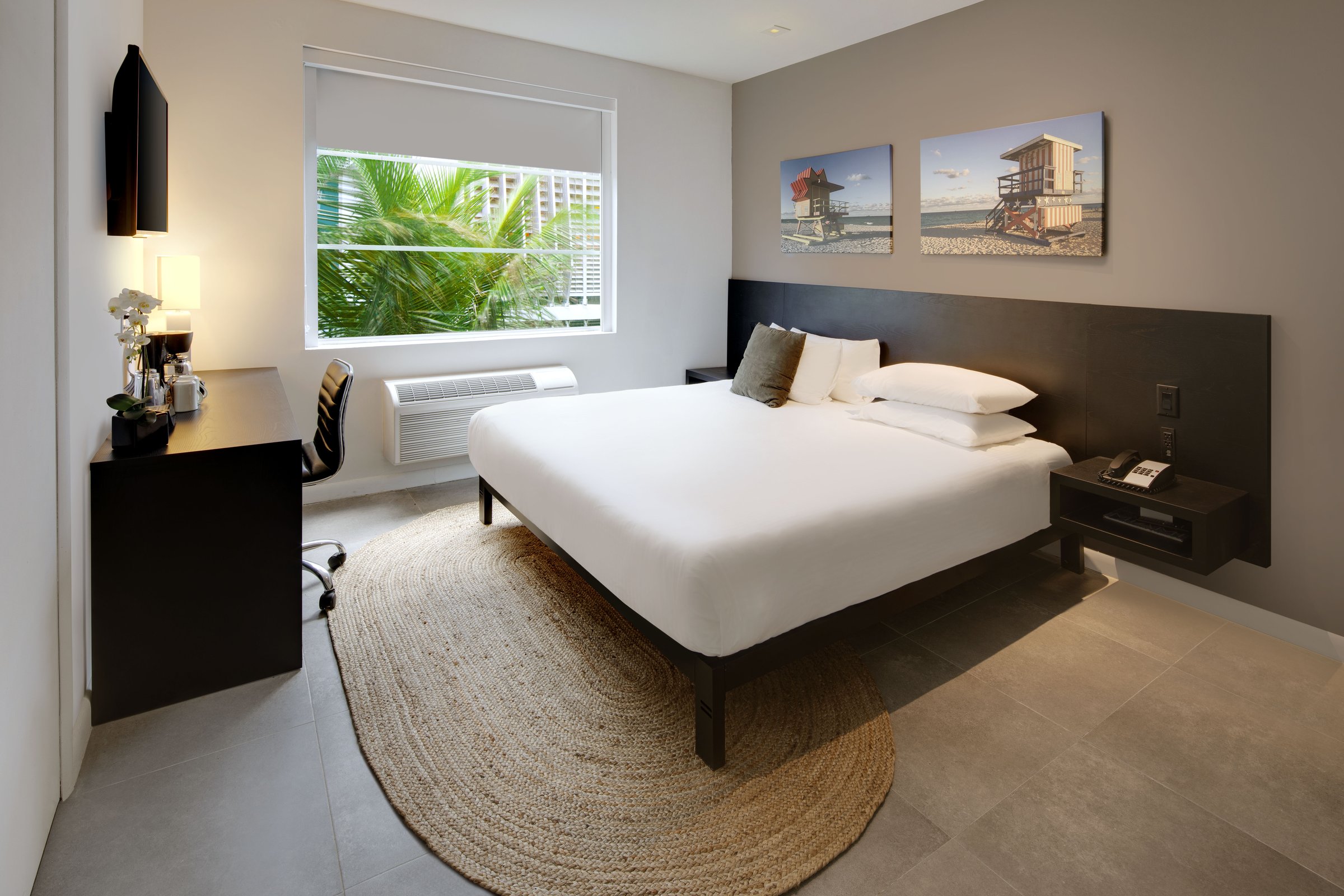 PNK1 Standard King, Accessible — bed and desk beneath twin lifeguard-tower canvases with an orchid on the desk and palm fronds at the window