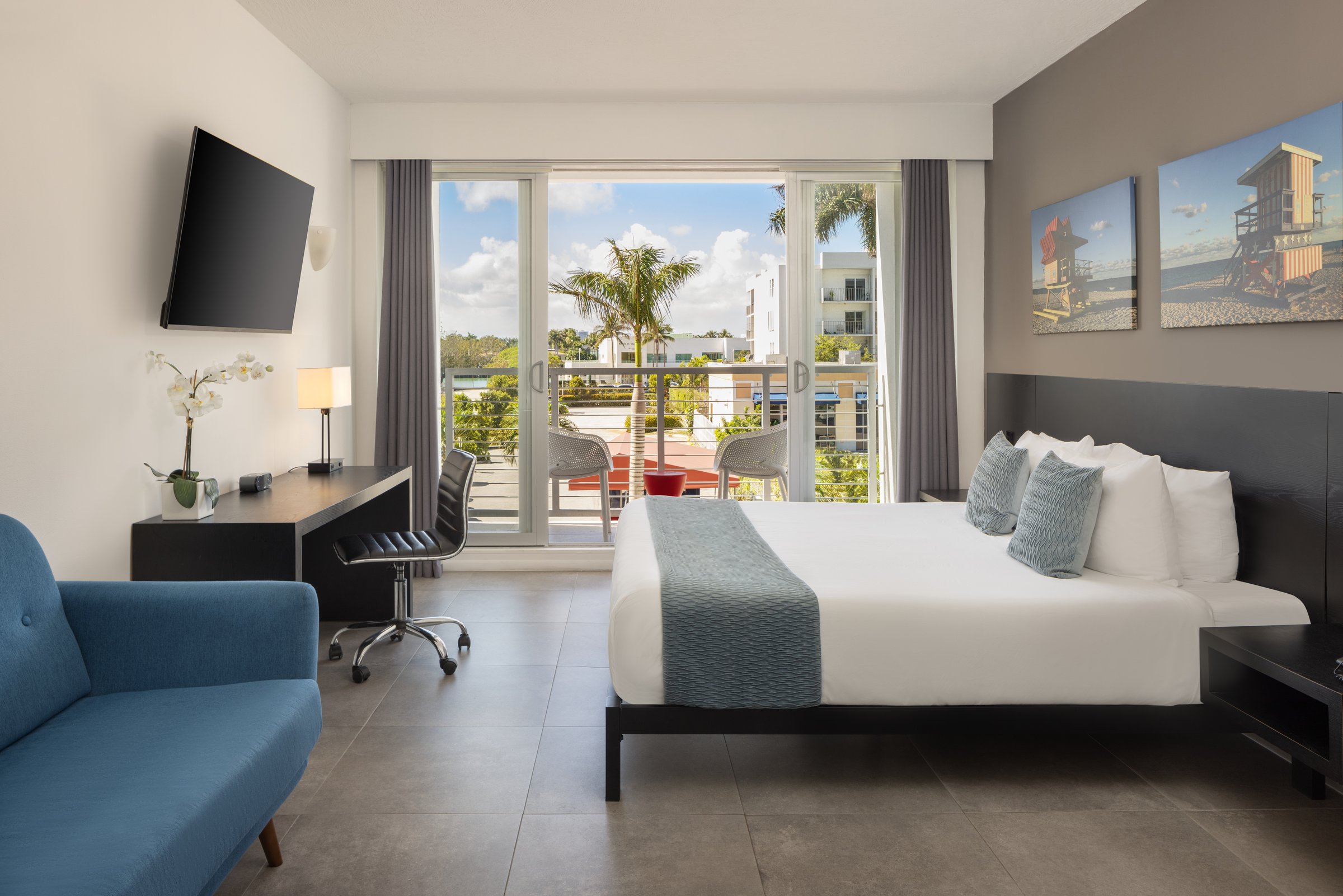 NK4 Superior King with City View Balcony — king bed with a slate runner facing sliding glass doors onto a balcony with palms and low rooftops beyond, blue sofa and desk to the left
