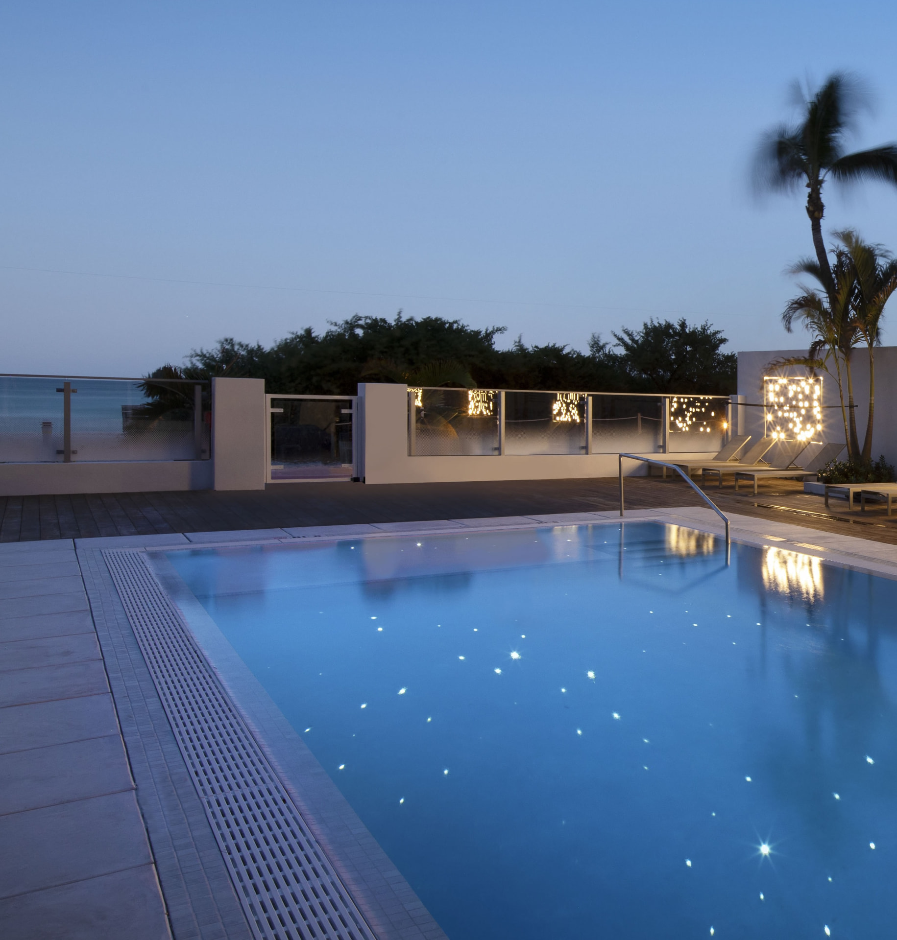 MB Hotel's pool deck at dusk, loungers arranged under palms with the illuminated mb monogram behind