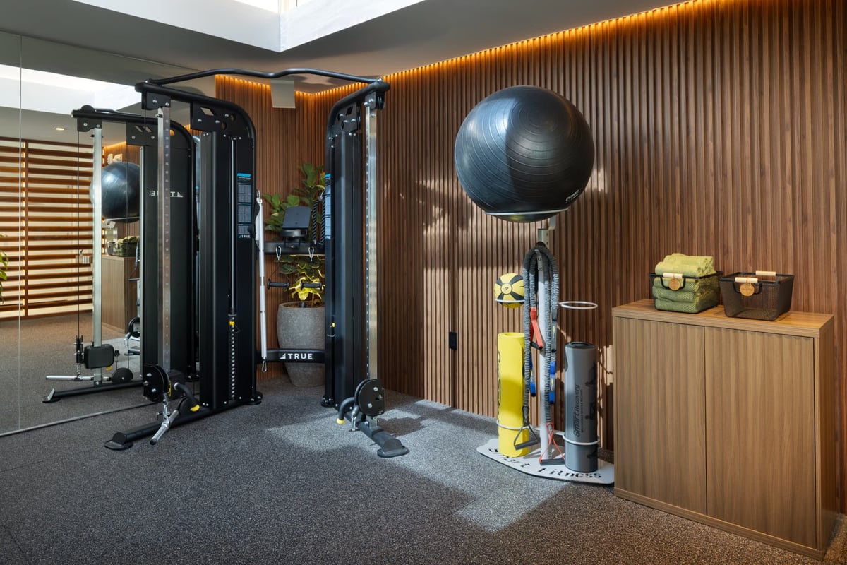 Functional trainer, stability ball and recovery tools against the backlit walnut wall of the MB Hotel gym, with folded towels on the cabinet