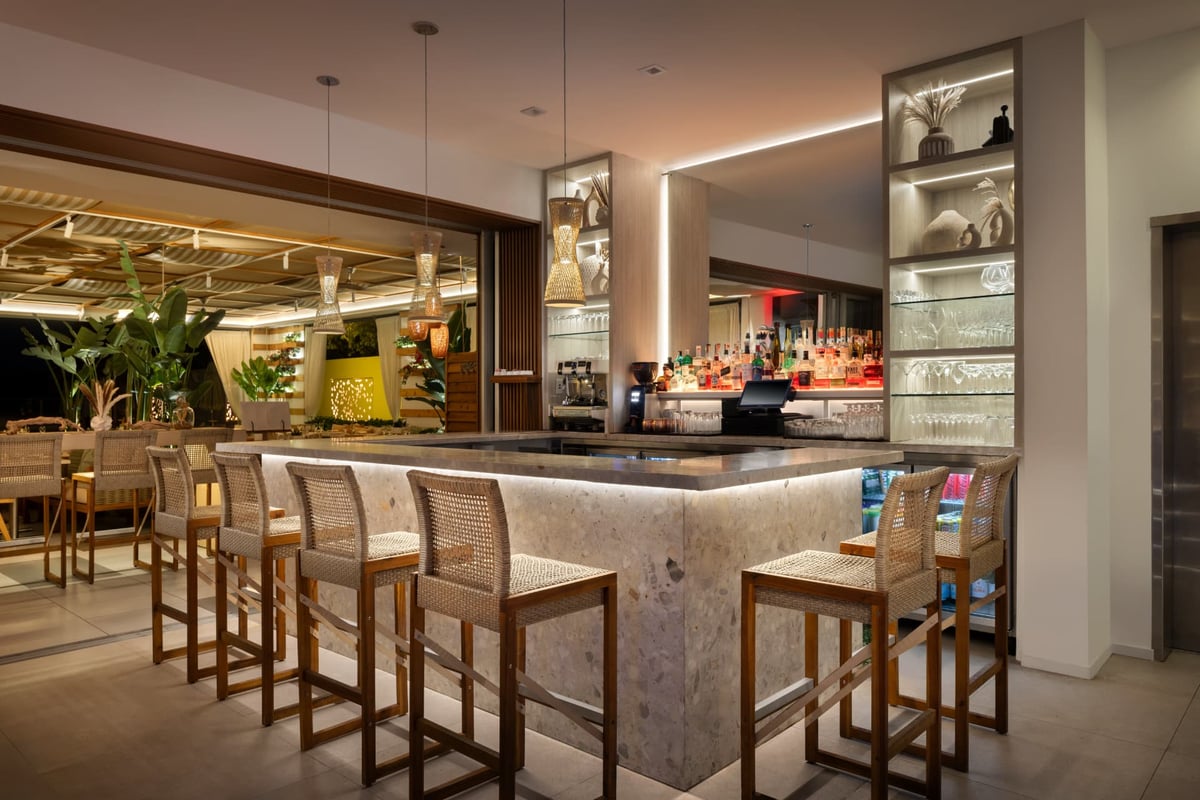 The lounge bar at MB Hotel after dark — a terrazzo counter lit from beneath, woven rattan pendants overhead and cane-back stools facing the illuminated garden terrace