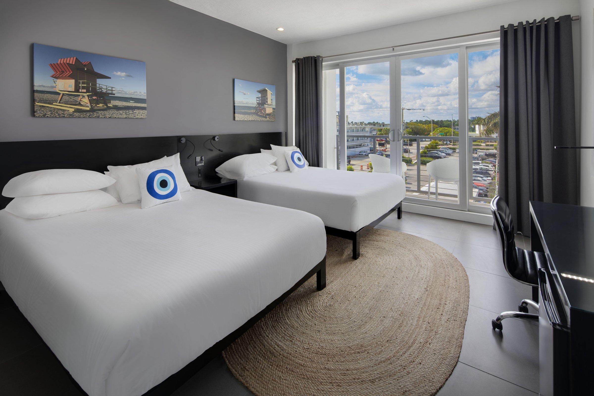 SNQ4 balcony outlook — beds in the foreground and the balcony doors open onto white balcony chairs above shopfronts and palms on Collins Avenue