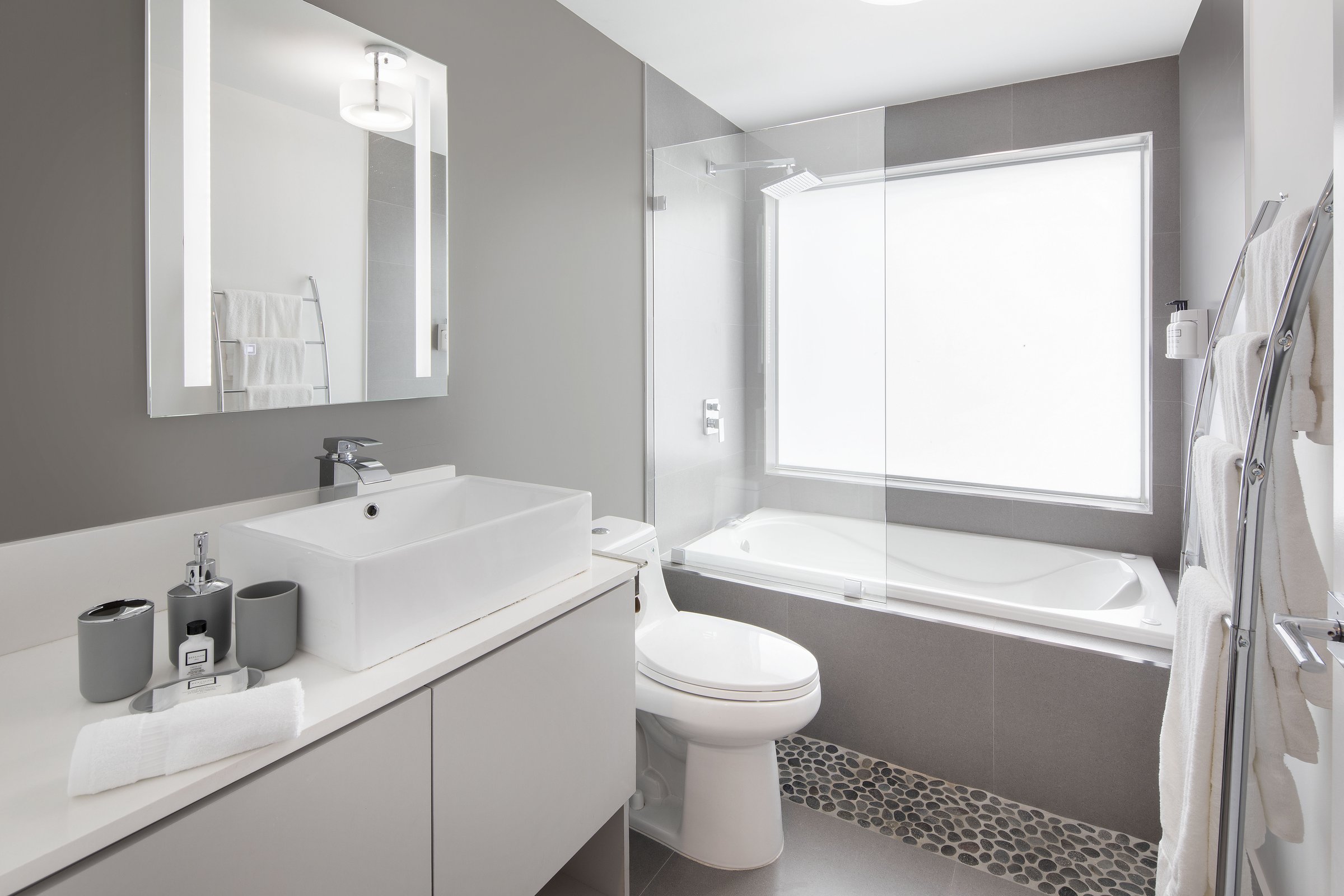 NQQ3 Superior Double Queen bathroom — white vessel basin on a pale vanity, glass-screened soaking tub with a frosted window above and a pebble-tile border at the floor