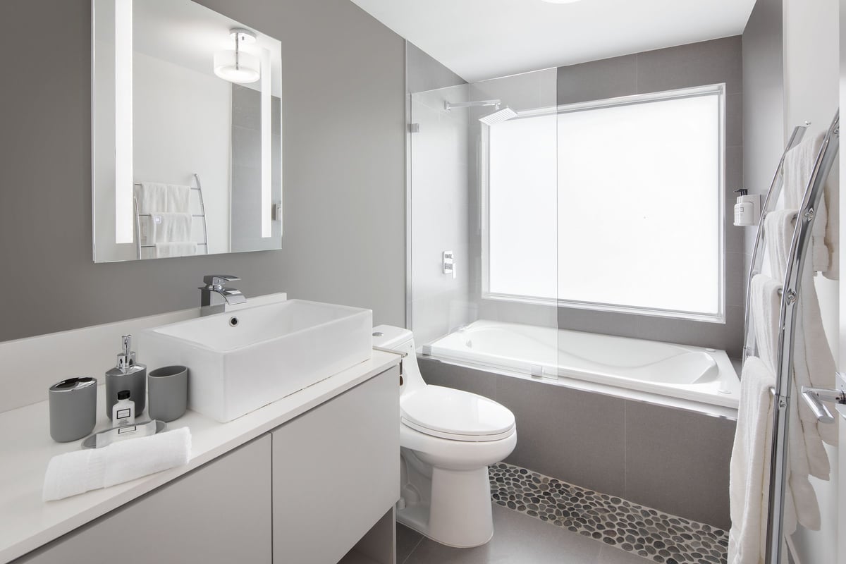 NQQ3 Superior Double Queen bathroom — white vessel basin on a pale vanity, glass-screened soaking tub with a frosted window above and a pebble-tile border at the floor