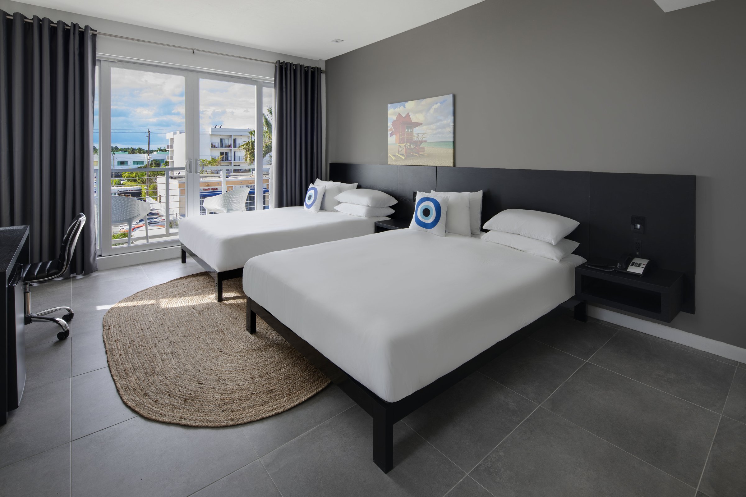 NQQ4 Deluxe Double Queen — beds seen from the desk wall with two lifeguard-tower canvases above the headboard and the balcony overlooking parked cars on the avenue