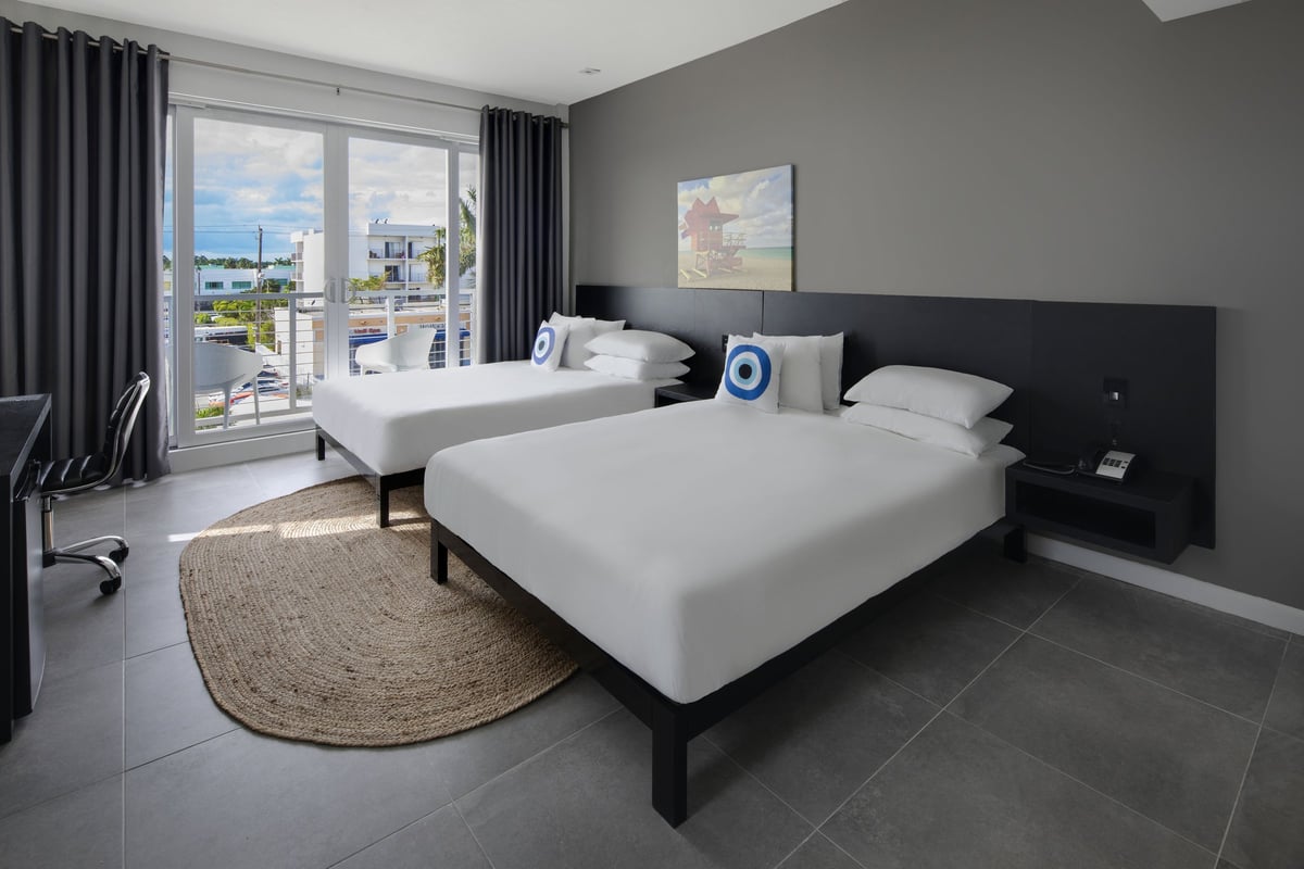 NQQ4 Deluxe Double Queen — beds seen from the desk wall with two lifeguard-tower canvases above the headboard and the balcony overlooking parked cars on the avenue