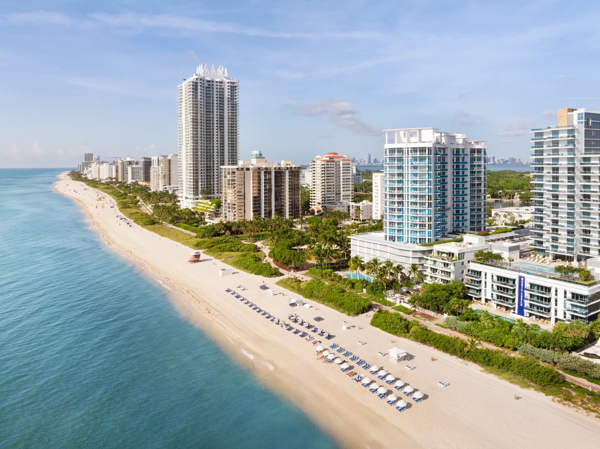 Aerial looking south along the Miami Beach coastline on a clear morning, turquoise water meeting a wide band of pale sand with the hotel towers and the downtown skyline in the distance