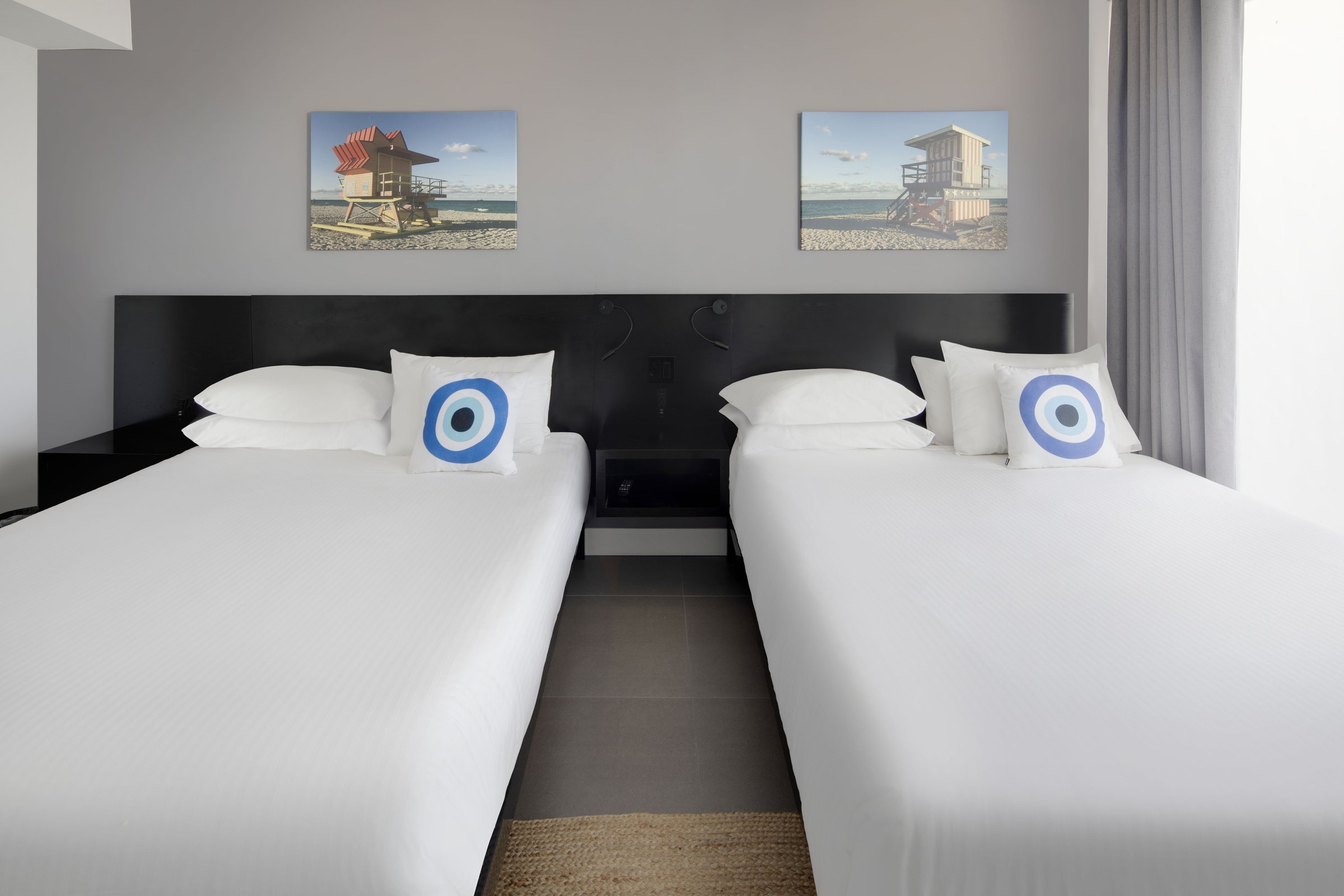SNQ4 bedroom — the two queen beds photographed head-on with blue evil-eye cushions and a pair of lifeguard-tower canvases above the shared black headboard