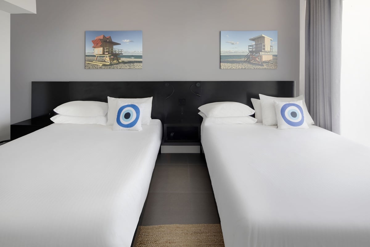 SNQ4 bedroom — the two queen beds photographed head-on with blue evil-eye cushions and a pair of lifeguard-tower canvases above the shared black headboard