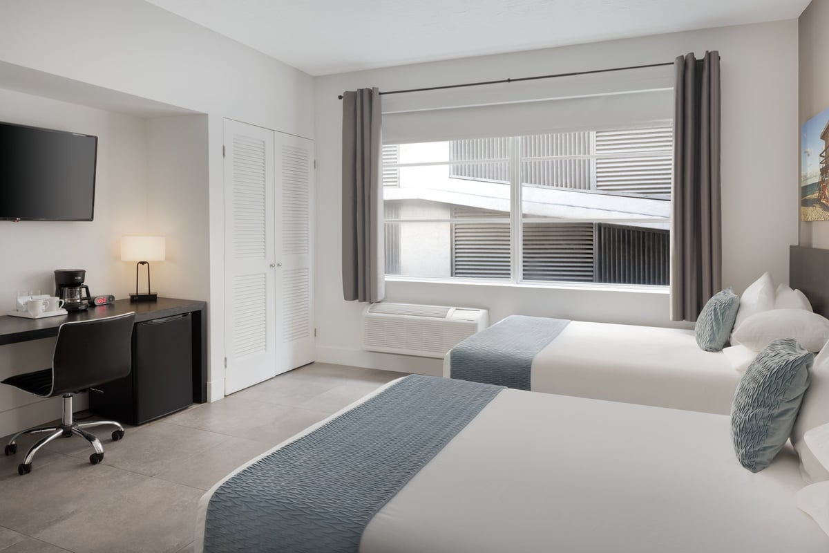 NDD1 Standard Double Full — two full beds with grey-blue bed runners beside a wide window, a black desk with coffee maker and cooler along the near wall