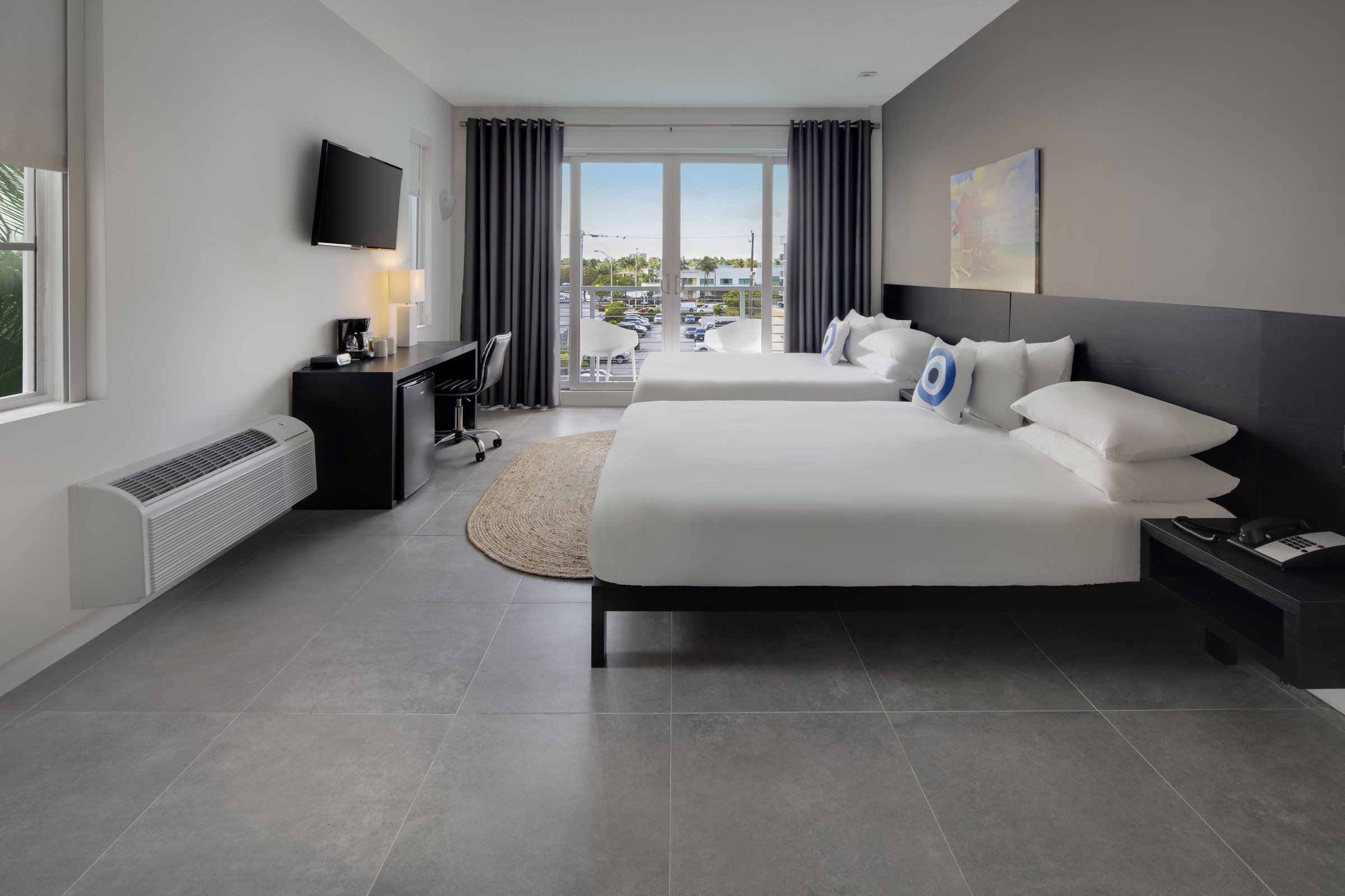 NQQ4 Deluxe Double Queen — both beds in plain white linen with blue evil-eye cushions, an oval jute rug between them and the balcony doors open to a bright sky