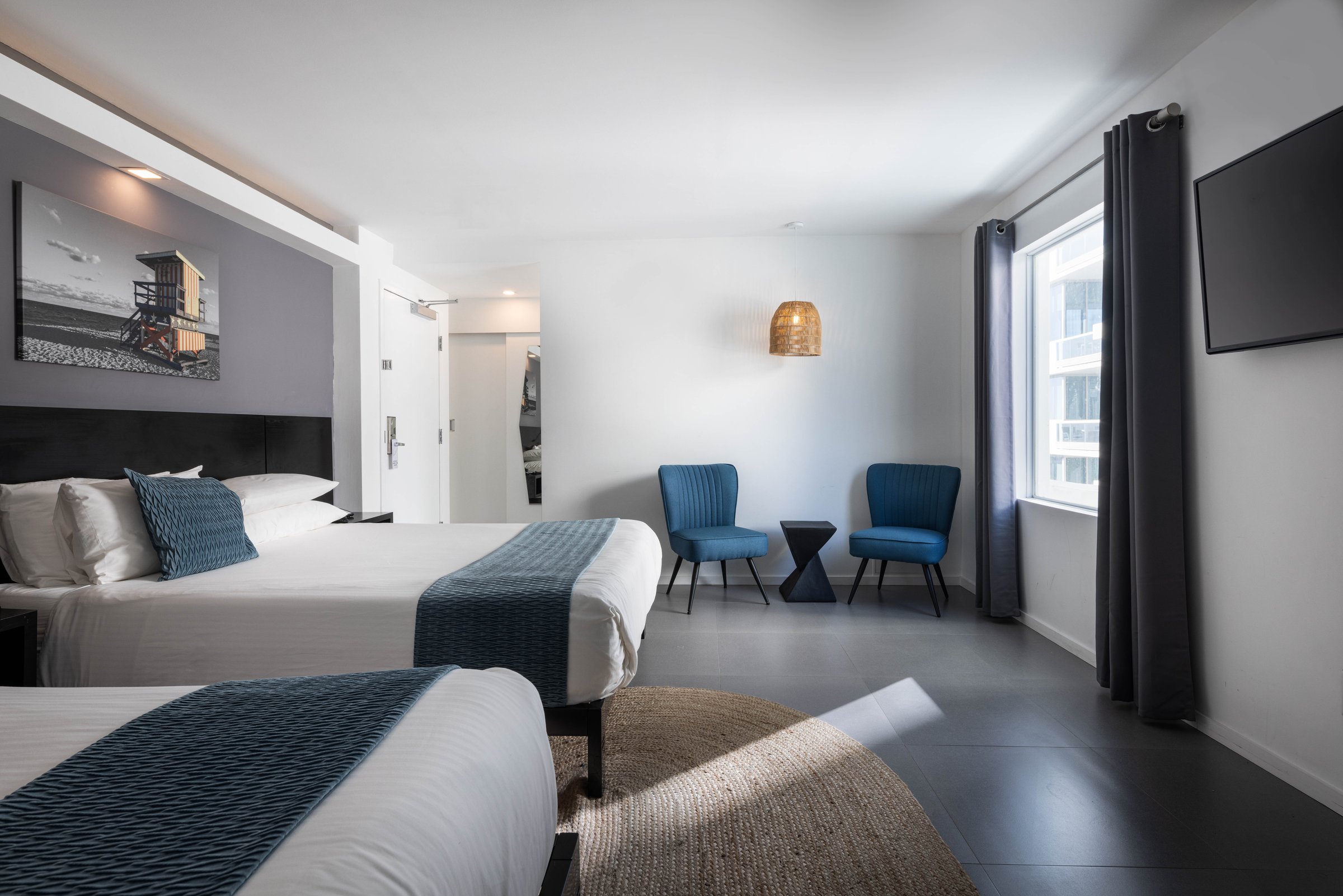 NQQ3 Superior Double Queen — two queen beds in white linen with blue evil-eye cushions beneath lifeguard-tower canvases, an oval jute rug and desk along the left wall